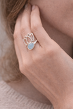 Bague en argent 925 avec pierre d’aigue-marine, ornée d’un motif fleur stylisée; portée à l’annulaire, vue sur la main et le visage partiel.