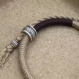 Bracelet Cercle Du Bonheur : cordon beige et noir avec fermoir argenté à motif, nœud et pendentif torsadé.